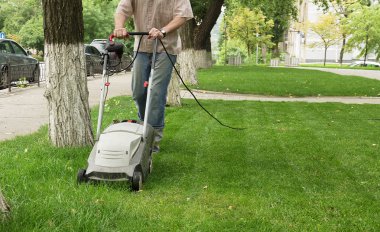 Worker with a electric lawn mower cares for an urban lawn near the sidewalk clipart