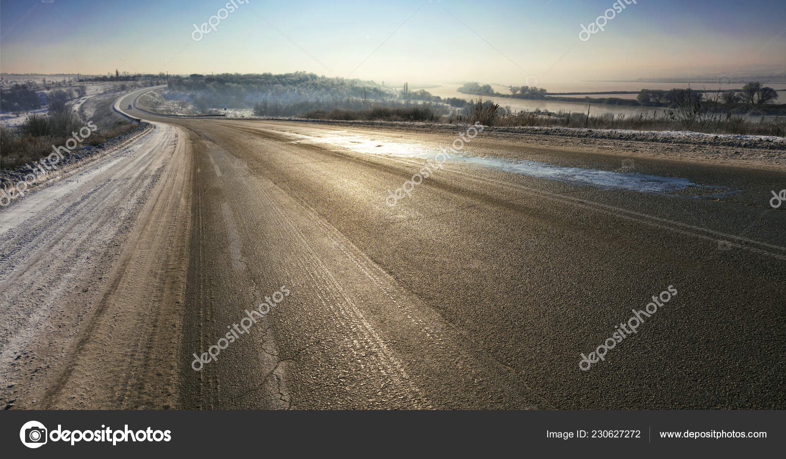 一条古老道路的柏油路在寒冷中裂开在蜿蜒的河流被白雪覆盖的田野和树木的背景下在蓝天的阴霾中被太阳的光线照亮 一条古老道路的柏油路在寒冷中裂开在蜿蜒的河流被白雪覆盖的田野和树木的背景下在蓝天的阴霾中被太阳的光线照亮