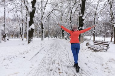 Genç güzel kadın parlak mercan ceket ve kot kış karla kaplı Ahşap banklar ile masal park avenue boyunca yürür ve kollarını geniş açılır