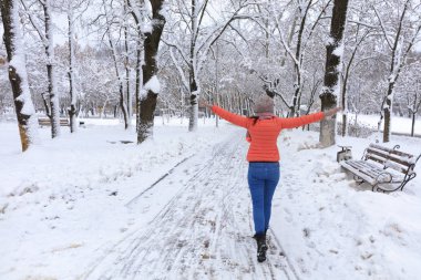 Genç güzel kadın parlak mercan ceket ve kot kış karla kaplı Ahşap banklar ile masal park avenue boyunca yürür ve kollarını geniş açılır