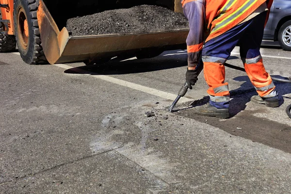 A road maintenance worker removes old asphalt on the roadway with a jackhammer into an excavator during road construction.