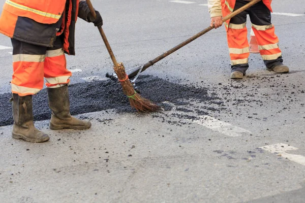 Two road builders assemble and level fresh asphalt with a whisk and level on the repaired part of the road.