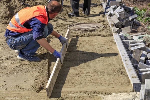 The worker clears and aligns the sand base with a wooden board for the subsequent laying of tiles on the sidewalk and around the sewer manhole.