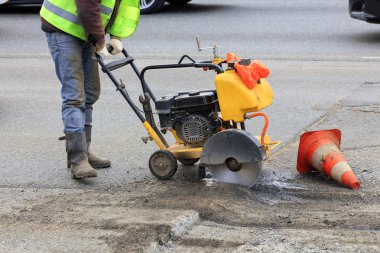 Worker cuts a piece of bad asphalt with a gasoline cutter during road construction. clipart