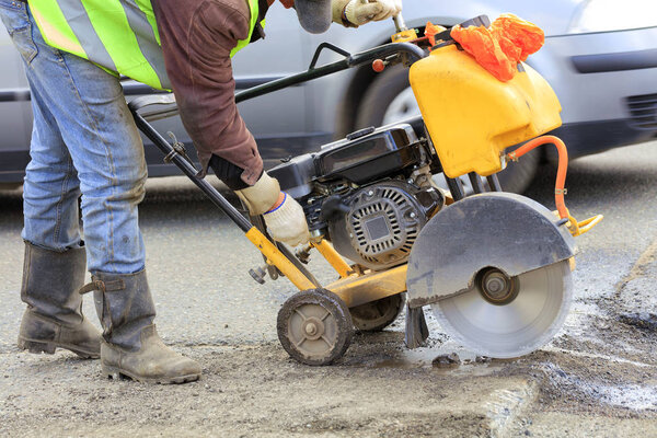 A worker in a green reflective vest starts the engine of a gasoline cutter to cut and clean bad asphalt on the roadway.