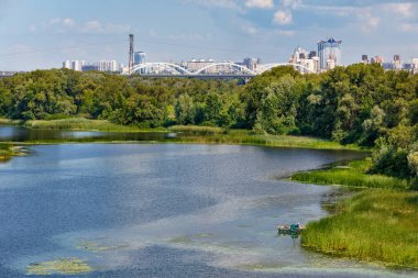 Dnipro 'nun güzel körfezi, kıyıya demirlemiş bir balıkçı teknesi ile kaplanmış bankalarla dolu ve ufukta şehir endüstrisi ve Kyiv' in manzarası var..