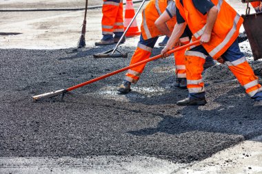 Turuncu tulumlu bir yol işçisi grubu, yolun bir bölümünü taze asfalt ile yeniliyor ve metal seviyesinde tamir edilmesi için düzleştiriyor..