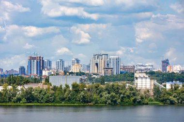 Dnipro Nehri ile Kyiv 'in sağ kıyısının manzarası ve ön planda bir endüstriyel kompleks ve sıcak bir yaz gününde ufukta yüksek binaları olan yeni bir yerleşim alanı manzarası..