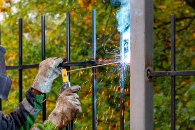 Koruyucu kasklı ve eldivenli bir kaynakçı, sonbahar yeşilliğinin bulanık arka planına karşı park alanındaki metal çitleri kaynak yapmak için elektrot kullanır. Seçici odak, boşluğu kopyala.