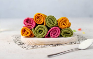 Sarı, pembe ve yeşil renkli krepler açık tahta bir tahtanın üzerinde. Beyaz bir masada, ekşi kremalı bir kaşığın yanında..