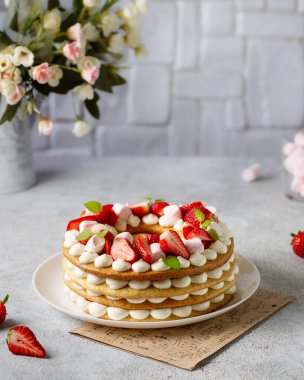 Krem şantili ve beyaz bir tabakta çilekli açık pasta. Tuğla bir duvarın ve bir buket çiçeğin arkasında.