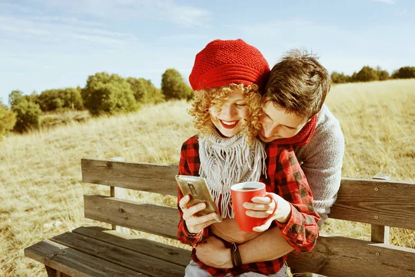 在秋天阳光明媚的日子里 幸福的年轻夫妇在草地上喝着咖啡 一边看着智能手机 一边拥抱和日光浴 — 图库照片