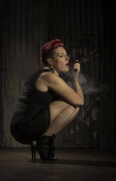 Sultry woman with fuchsia hair crouching and holding a cigar