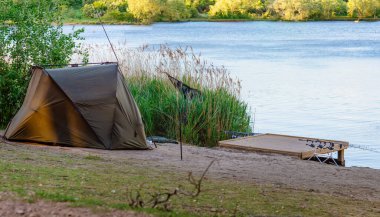Gün batımında göl kıyısında bir çadır ve tatlı suda sazan balığı avlama çubuğu.