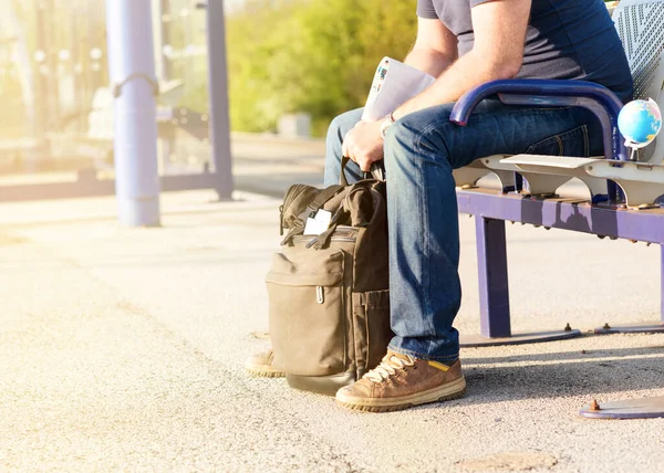 Man waiting train with a green travel backpack, maps, glasses on the platform at the train station at the sunset