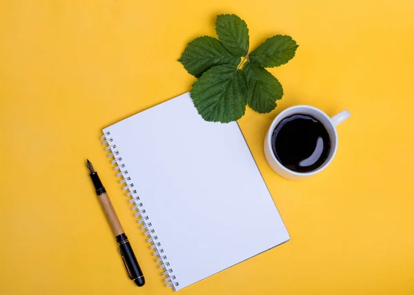 A cup of coffee,  empty paper, pen and leaf on yellow background