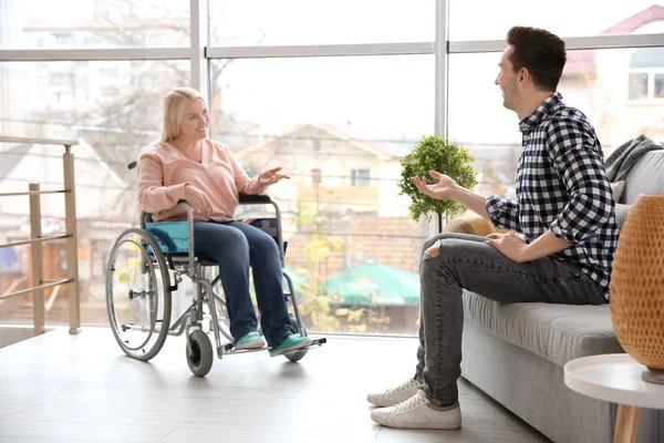 Hombre joven hablando con mujer madura en silla de ruedas en interiores — Foto de Stock