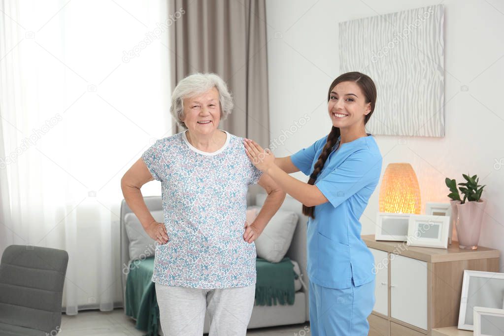 Senior woman doing therapeutic gymnastics with assistance of young caregiver indoors
