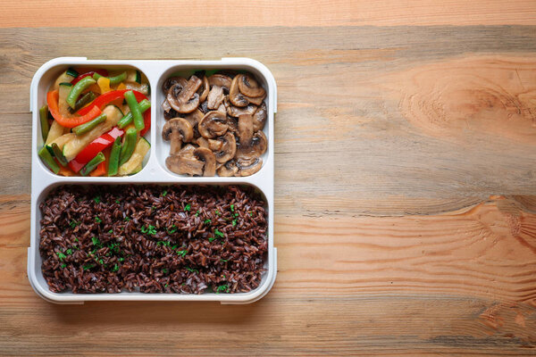 Lunch box with red rice, vegetables and mushrooms on table