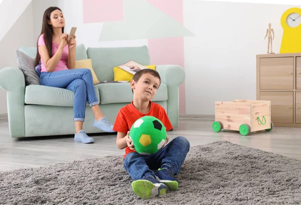 Kleiner Junge spielt zu Hause mit Ball, während sein Kindermädchen Handy benutzt — Stockfoto