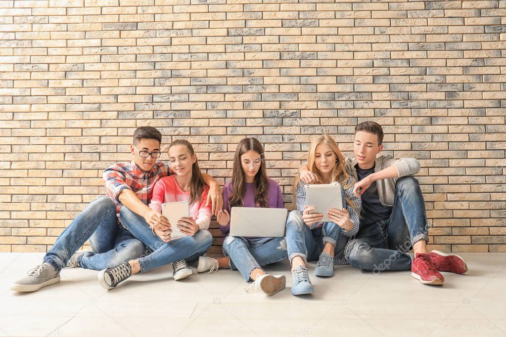 Teenagers with modern devices sitting on floor near wall