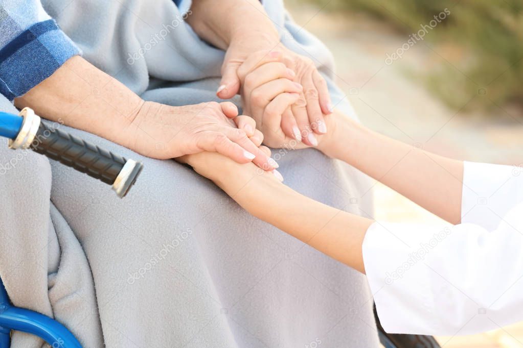 Young nurse from care home holding hands of senior woman in wheelchair outdoors, closeup