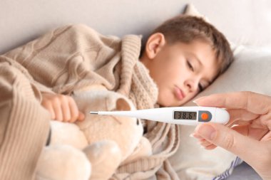 Woman holding thermometer with high temperature and sick child lying on couch at home clipart