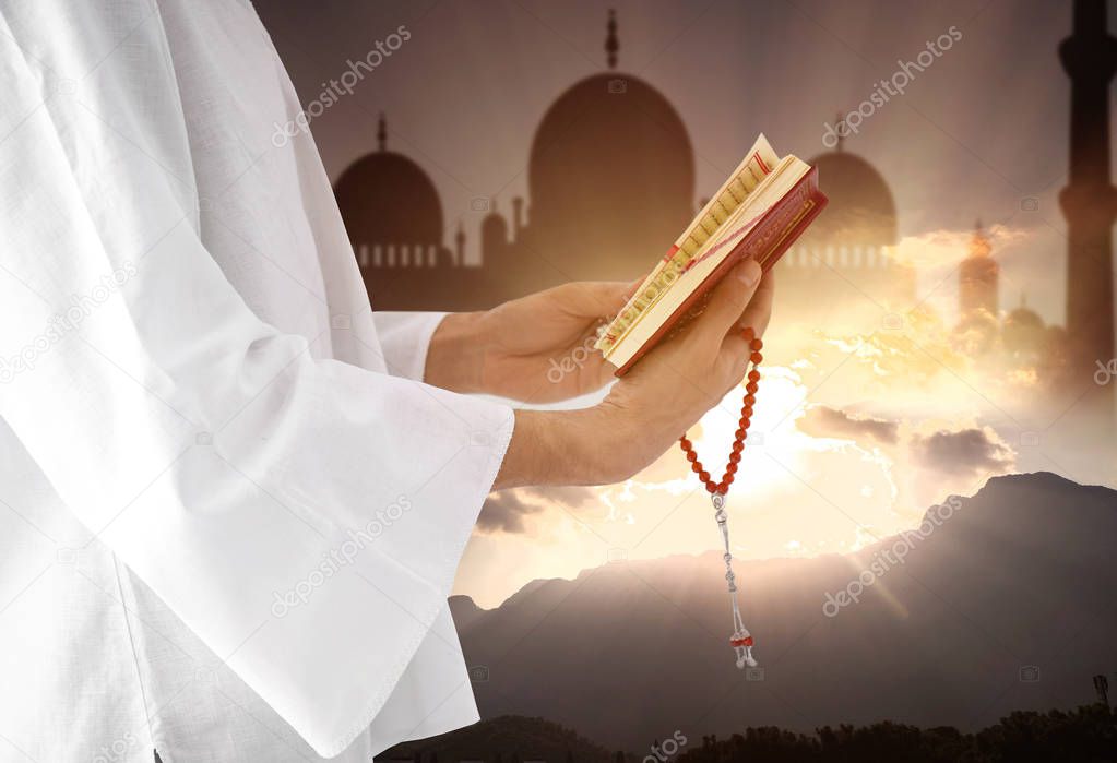 Young Muslim man reading Koran and silhouette of mosque with landscape on background