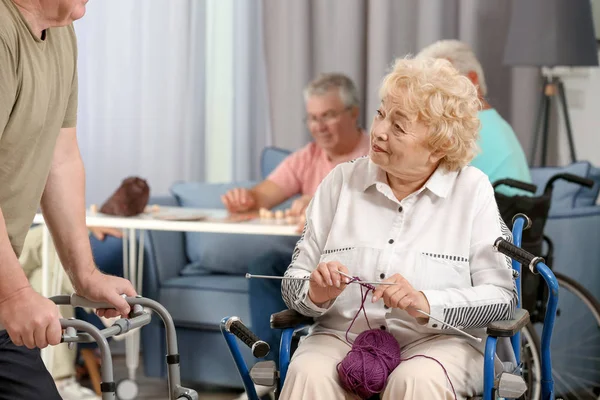 Mujer mayor en silla de ruedas tejiendo mientras habla con su amigo en el hogar de cuidado —  Fotos de Stock