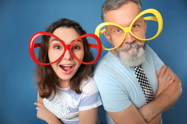 Ung kvinna och mogen man i roliga glasögon på färgbakgrund. April fool's day firande — Stockfoto
