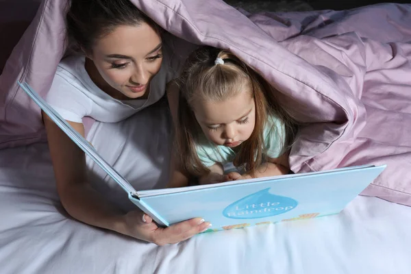 Ibu membaca cerita pengantar tidur untuk putri kecil saat berbaring di bawah selimut — Stok Foto