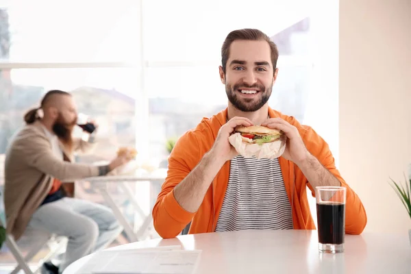 Красивый мужчина ест вкусный бургер в кафе — стоковое фото
