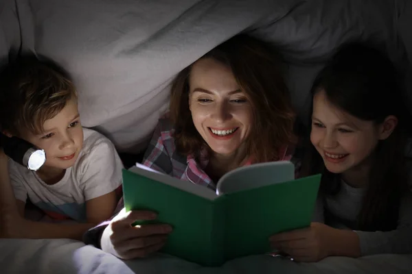 Mother reading bedtime story to her little children under blanket in evening