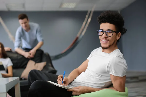 African American man writing in notebook while other students resting together at campus building — Stock Photo, Image