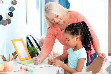 Female mature nanny helping little African-American girl to do homework clipart