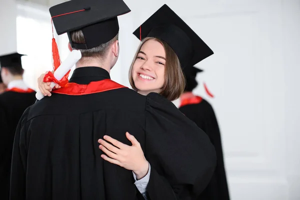 Estudantes felizes em roupões de solteiro abraçando dentro de casa, dia da formatura — Fotografia de Stock