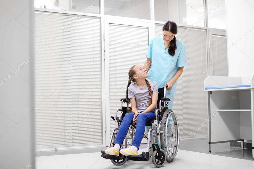 Young female doctor taking care of little girl in wheelchair indoors