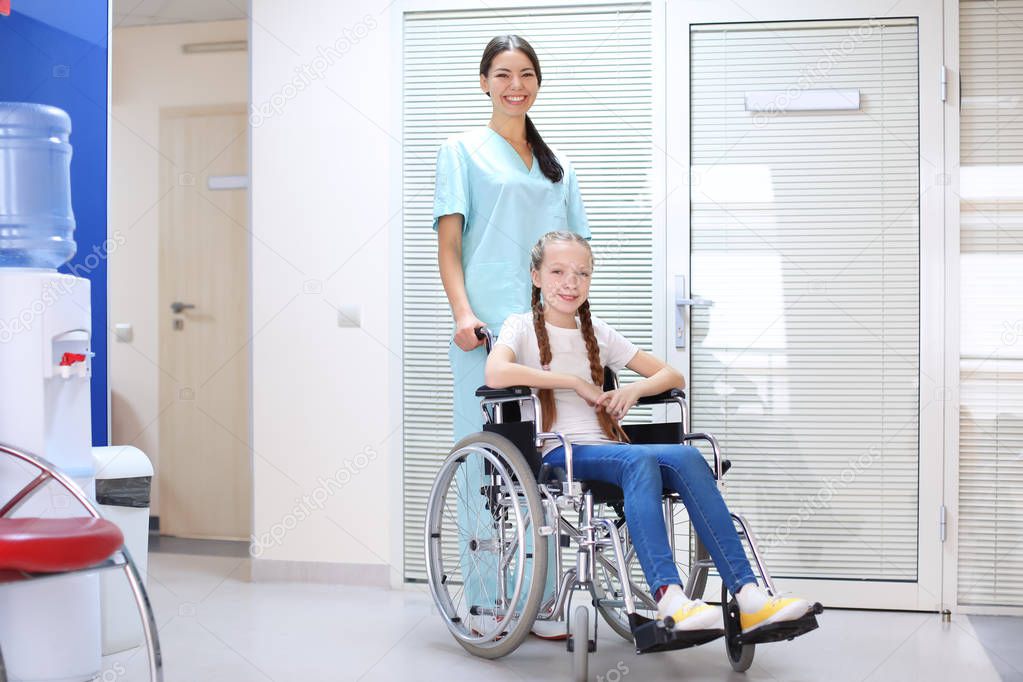 Young female doctor taking care of little girl in wheelchair indoors