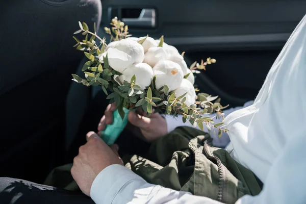 Wedding bouquet of white peony in the hands of the groom sitting in the car, selective focus