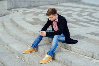 Şık genç ekose gömlek ve kot merdivenlerde oturan adam ve açık havada, smartphone kullanarak tam vücut. Öğrenci ile smartphone, boş alan