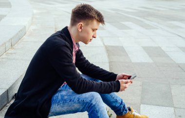 Ekose gömlek ve merdivenlerde oturan ve açık havada smartphone kullanarak kot şık genç adam. Öğrenci ile smartphone, boş alan