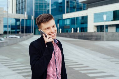 Mutlu genç adam ekose gömlek ve şehirde yürüyüş ve cep telefonu açık havada üzerinde söz ceket. Öğrenci ile hareket eden telefon