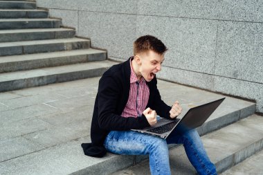 Dizüstü bilgisayar ile mutlu bir adam. Başardım! Genç adam bilgisayar monitörü dizlerinin üzerinde seyir ve Not. Görev ve yumruk açık havada sıktı elleri ile zafer mutlu adam tamamlandı