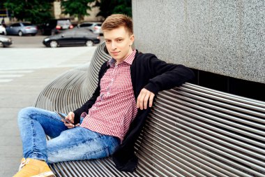 Ekose gömlek ve ceket ile cep telefonu bankta el açık havada oturup şık rahat genç adam portresi. Öğrenci seyahat