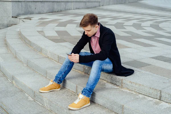 Şık genç ekose gömlek ve kot merdivenlerde oturan adam ve açık havada, smartphone kullanarak tam vücut. Öğrenci ile smartphone, boş alan