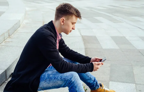 Ekose gömlek ve merdivenlerde oturan ve açık havada smartphone kullanarak kot şık genç adam. Öğrenci ile smartphone, boş alan