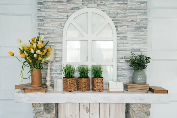 White wooden shelf with bouquet of tulips, grass in pot, greenery in vase, books and candle against stone wall with decorative window. lose-up decoration in living room at home