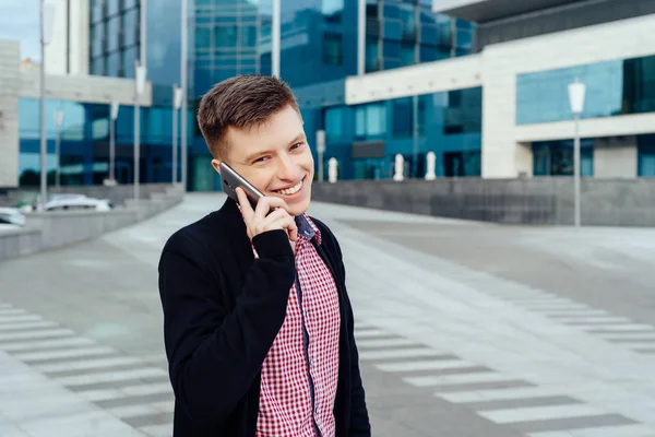 Mutlu genç adam ekose gömlek ve şehirde yürüyüş ve cep telefonu açık havada üzerinde söz ceket. Öğrenci ile hareket eden telefon