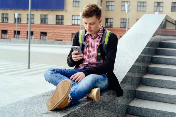 Taş merdiven yanında oturan ve akıllı telefon şehir açık, tam vücut kullanarak sırt çantası ile mutlu yakışıklı genç adam. Öğrenci üniversite açık havada sonra aittir