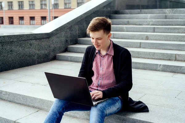 Merdivenlerde oturan ve açık havada dizüstü bilgisayarda yazarak kot pantolon ve ceket şık genç adam. Teknoloji ve iletişim kavramı 